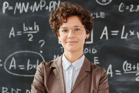 Lächelnde Lehrerin mit Brille und lockigem Haar steht vor einer Tafel mit mathematischen Formeln