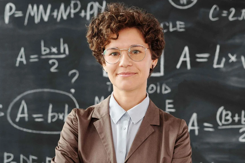Lächelnde Lehrerin mit Brille und lockigem Haar steht vor einer Tafel mit mathematischen Formeln